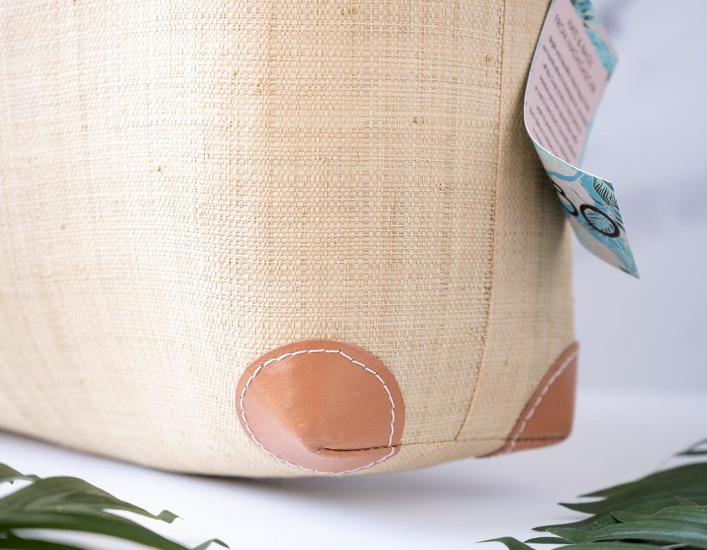 Closeup View of the Bottom Corner of the End View of the Shebobo Bebe Straw Bag with Bamboo Handles against a clean white and marble background with palm leaves