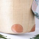 Closeup View of the Bottom Corner of the End View of the Shebobo Bebe Straw Bag with Bamboo Handles against a clean white and marble background with palm leaves