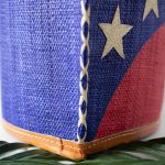 Close Up Side View of the American Flag Raffia Tote Bag by Shebobo Against a Marble and Clean White Background with Palm Leaves