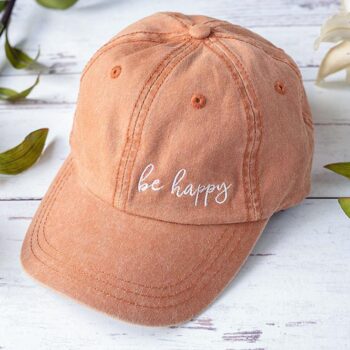 Top View of the Orange Women's Be Happy hat sitting on a white rustic wood background between 2 flowers and stems