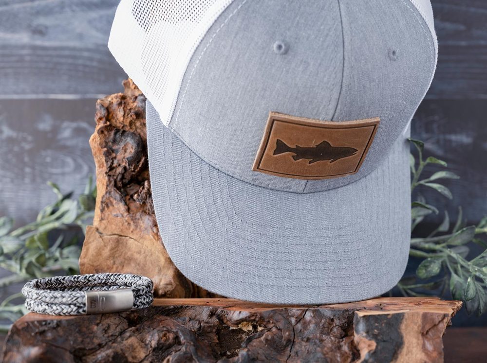 Overcast Waters Perfect Pairing of the Trout Hat by RANGE LEather and Rope Lake Bracelet by Steel & Barnett on a Piece of Wood with Rustic Background and Greenery