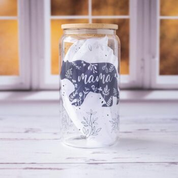 Front view of the Mama Bear glass tumbler with a mama bear and floral pattern on a rustic background with kitchen window