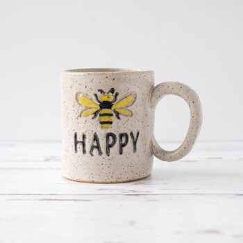 White Squirrel Clayworks stoneware ceramic bee happy coffee mug on white top with rustic white wood background