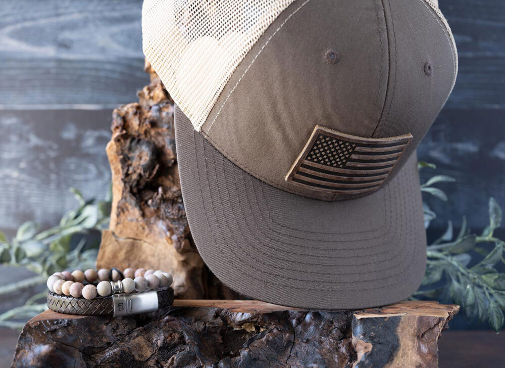 New Beginnings Sunsationsal Set Collection of the Brown & Khaki American Flag Leather Patch Hat with the Steel & Barnett Rainbow Beaded Bracelet and Cornall Leather Bracelet in Vintage Black on Wood and Against a Rustic Background