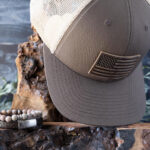 New Beginnings Sunsationsal Set Collection of the Brown & Khaki American Flag Leather Patch Hat with the Steel & Barnett Rainbow Beaded Bracelet and Cornall Leather Bracelet in Vintage Black on Wood and Against a Rustic Background