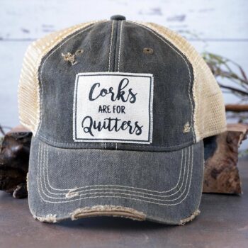 Corks are for quitters distressed trucker cap by Vintage Life on a rustic piece of wood and backdrop with greenery behind