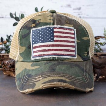 Women's Camouflage American Flag distressed trucker hat against a rustic background and greenery