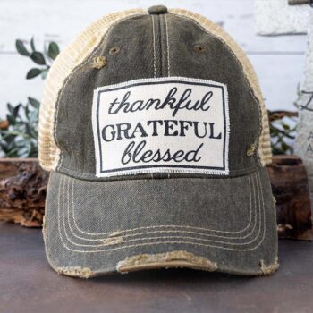 Women's thankful, grate, blessed distreesed trucker hat against a rustic background and greenery