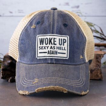 Woke up Sexy as Hell Again distressed trucker cap by Vintage Life on a rustic piece of wood and backdrop with greenery behind