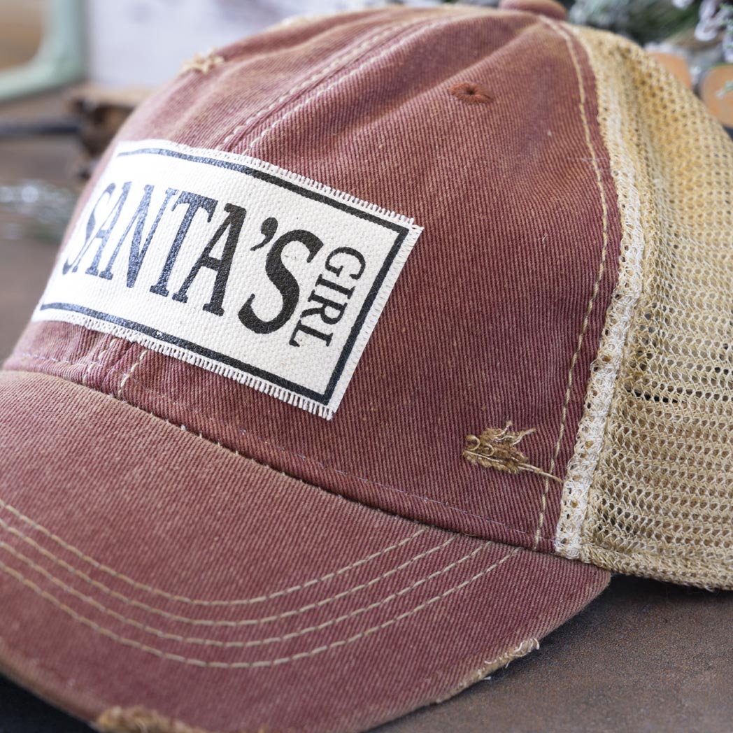 Angled view of the Vintage Life's Santa's Girl distressed trucker cap in turquoise and against a rustic background