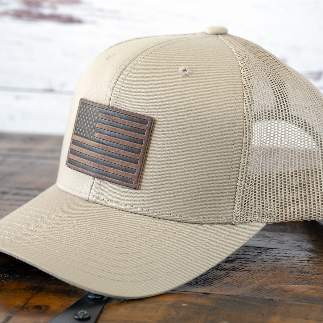 Left angle of the RANGE Leather American flag leather patch hat in tan against a rustic wood background