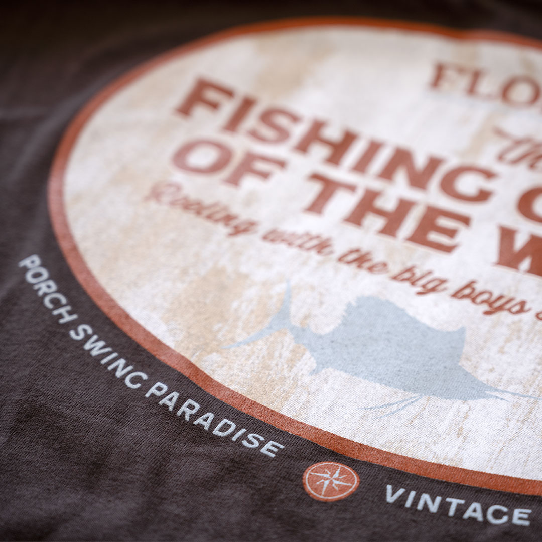 Closeup view of the back graphic lower on the Porch Swing Paradise Graphic Tee Florida... The Fishing Capital of the World graphic tee in the color chocolate against a rustic backdrop