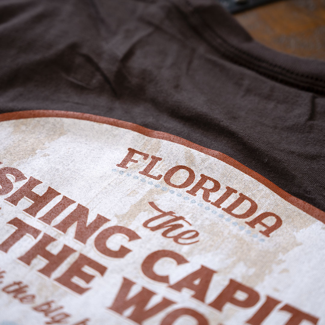 Closeup view of the back graphic top on the Porch Swing Paradise Graphic Tee Florida... The Fishing Capital of the World graphic tee in the color chocolate against a rustic backdrop
