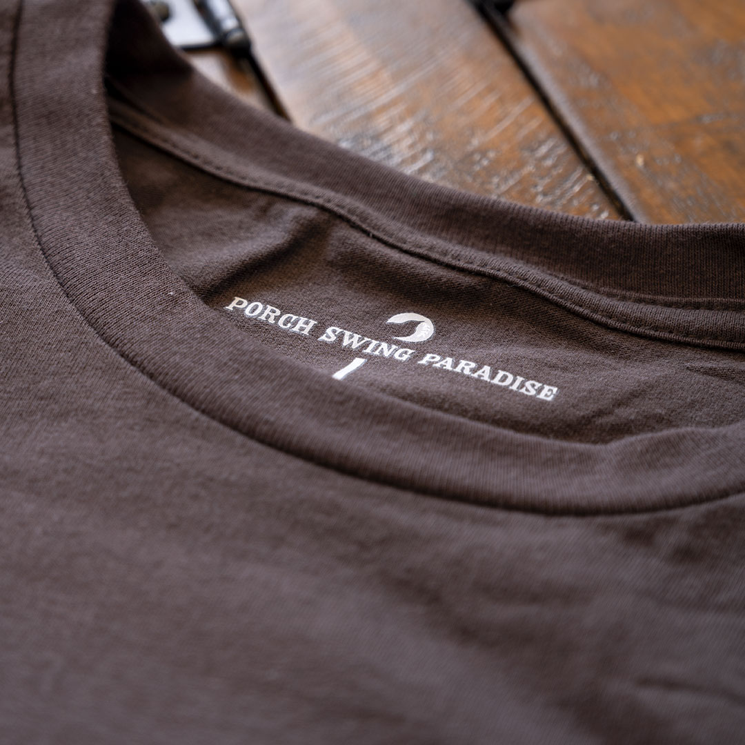 Closeup view of the branded inside tagging on Porch Swing paradise Graphic Tee Florida... The Fishing Capital of the World graphic tee in the color chocolate against a rustic backdrop