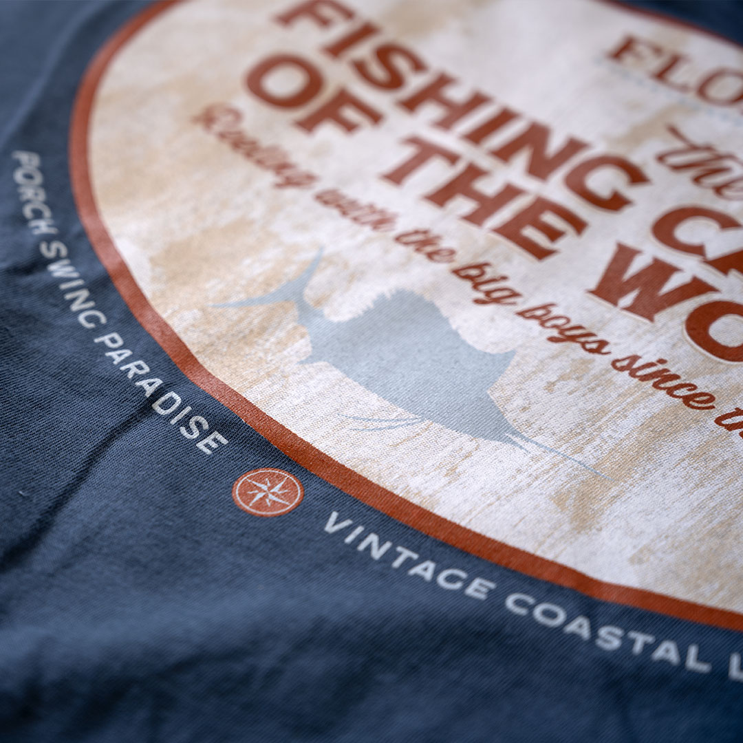 Closeup view of the back graphic lower on the Porch Swing Paradise Graphic Tee Florida... The Fishing Capital of the World graphic tee in the color indigo against a rustic backdrop