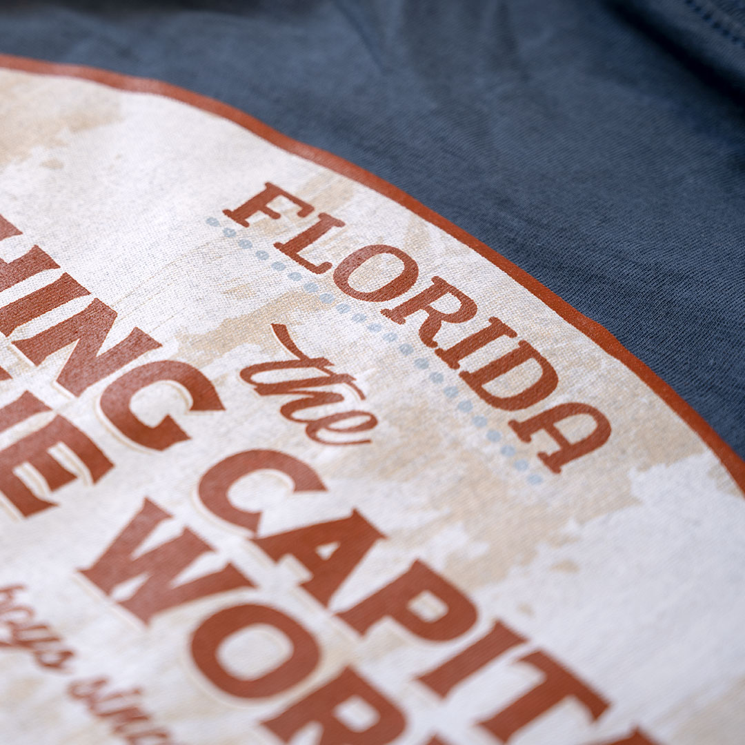 Closeup view of the back graphic top on the Porch Swing Paradise Graphic Tee Florida... The Fishing Capital of the World graphic tee in the color indigo against a rustic backdrop