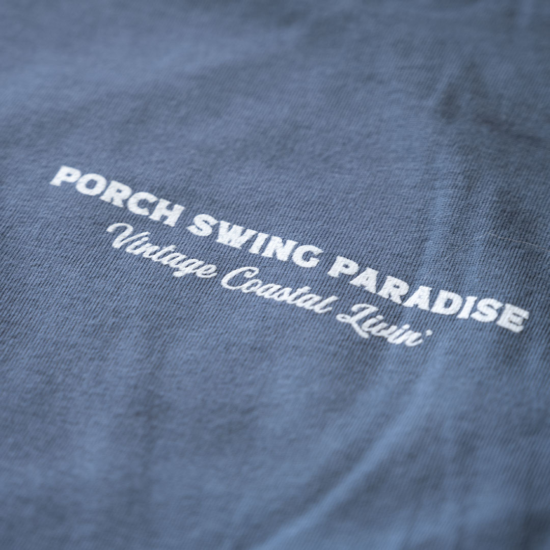 Closeup view of the front chest branded Porch Swing Paradise Graphic Tee Florida... The Fishing Capital of the World graphic tee in the color indigo against a rustic backdrop