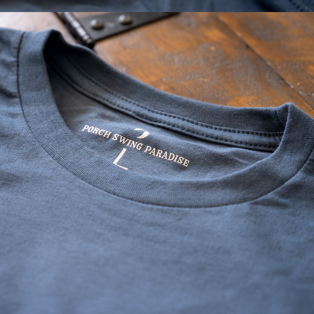 Closeup view of the branded inside tagging on Porch Swing Paradise Graphic Tee Florida... The Fishing Capital of the World graphic tee in the color indigo against a rustic backdrop