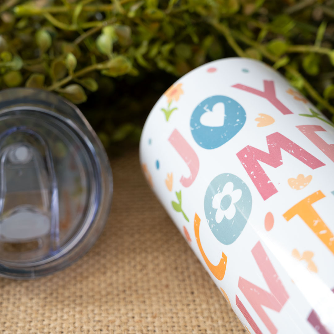 Closeup view of the Joy Comes in the Morning stainless skinny tumbler against greenery and with the top open and bamboo lid removed next to it
