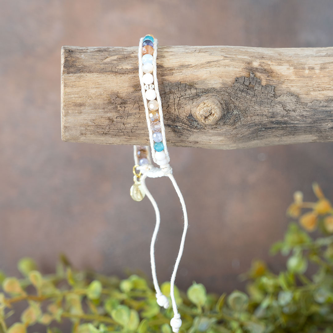 The Coastal Sunset Moonbeam Bracelet dangling on a piece of driftwood with a rustic backdrop