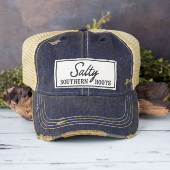 Salty Southern Roots distressed trucker cap in the color navy laying against driftwood and in front of greenery with a rustic backdrop