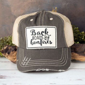 Backroads & Bonfires distressed trucker cap in the color dark gray laying against driftwood and in front of greenery with a rustic backdrop