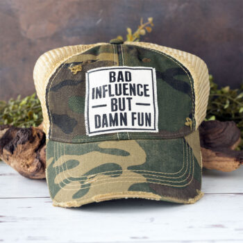 Bad Influence but Damn Fun distressed trucker cap in the color camo laying against driftwood and in front of greenery with a rustic backdrop
