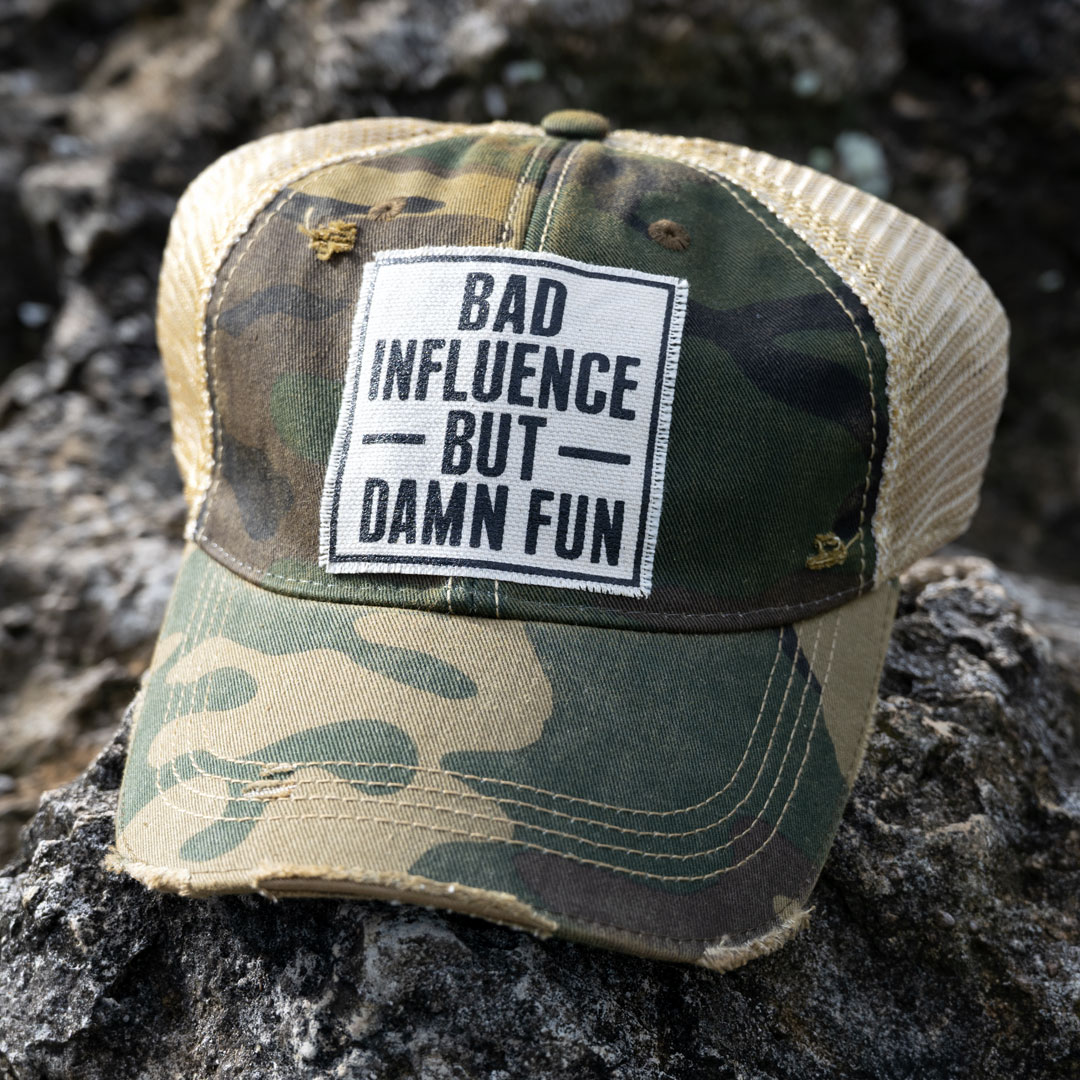 Bad Influence but Damn Fun distressed trucker cap in the color camo laying on a rock in the backroads near the bay
