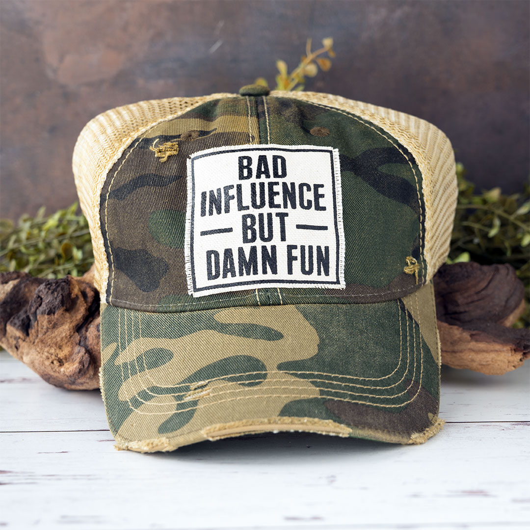 Bad Influence but Damn Fun distressed trucker cap in the color camo laying against driftwood and in front of greenery with a rustic backdrop