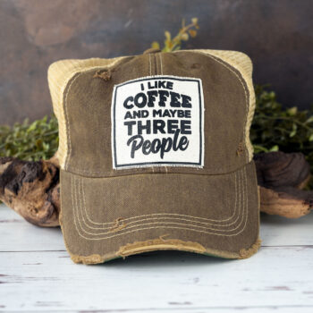 I Like Coffee and Maybe Three People distressed trucker cap in the color brown laying against driftwood and in front of greenery with a rustic backdrop