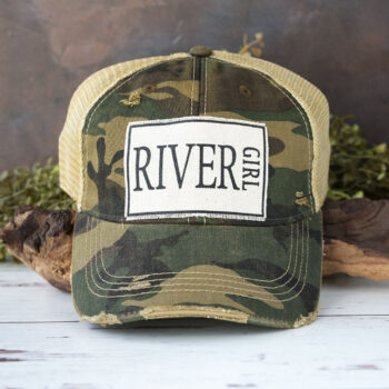 River Girl distressed trucker cap in the color camo laying against driftwood and in front of greenery with a rustic backdrop