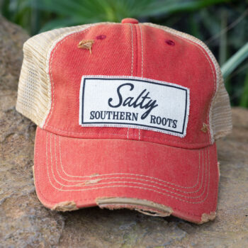 Salty Southern Roots distressed trucker cap in the color red laying in a big rock in the backwoods